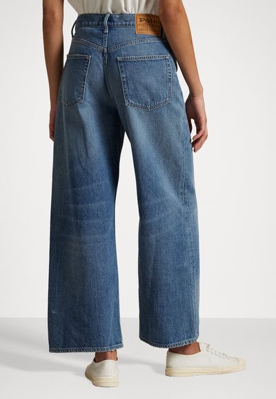 Vide ben, lyse blå denimbukser med baklommer; har en sydd logo-patch i midjen. Myk tekstur og avslappet passform.