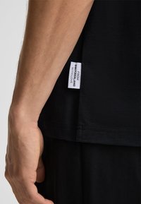 Schwarzes Baumwoll-T-Shirt mit geradem Saum, das an der Seitennaht ein weißes Stoffetikett trägt mit der Aufschrift "AUS DER SCHWEIZ MIT LIEBE".