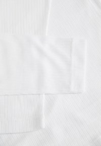 Haut blanc à manches longues côtelées, étalé à plat, montrant la texture et le détail du poignet sur un fond blanc uni.