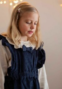 Blå kordflöjelklänning med volangdetaljer och en rosett, lagd över en vit långärmad tröja; ljusblont hår med en stjärnformad klämma.