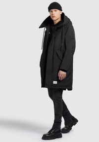 Svart vadderad parkas med stor luva, dragsnören, framfickor och en logotypmärke. Bärs över en svart hoodie och jeans med klumpiga svarta stövlar.