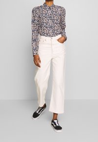 Camicia floreale a maniche lunghe con sfondo scuro, fiori bianchi e blu; abbinata a jeans bianchi a gamba larga e alta vita e sneakers nere.