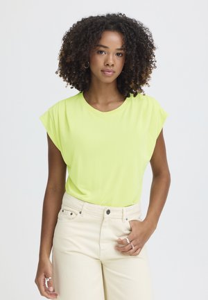 Ung kvinde med krøllet hår iført en lysegrøn, ærmeløs top og cremefarvede bukser med høj talje, stående med den ene hånd i lommen.