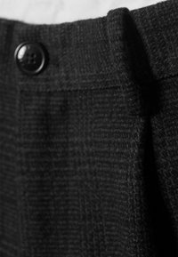 Črn teksturiran material hlač, ki ima ravno sprednjo stran, naborke in okroglo črno gumb na pasu. Detajlni posnetek.