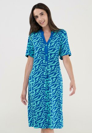 Mujer con un vestido hasta la rodilla de color azul y turquesa con estampado, mangas cortas y botones delanteros, posando contra un fondo liso.