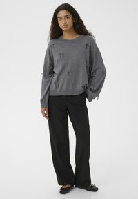 Ženska z dolgimi kodrastimi lasmi, oblečena v sivi pulover z majhnimi pentljicami in črne široke hlače, stoji pred enostavnim ozadjem.