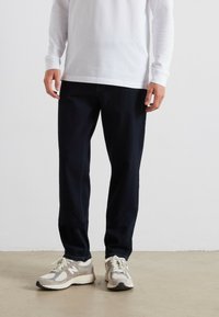 Jeans in denim blu scuro con taglio dritto, abbinati a sneaker grigie e crema. La maglietta a maniche lunghe è bianca e presenta un collo a giro.