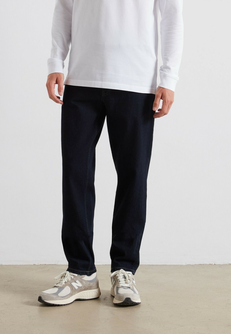 Jeans in denim blu scuro con taglio dritto, abbinati a sneaker grigie e crema. La maglietta a maniche lunghe è bianca e presenta un collo a giro.