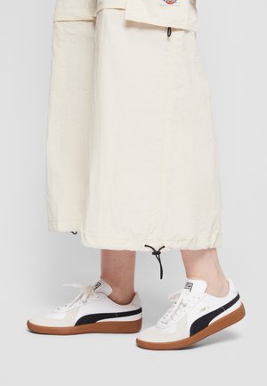 Białe sneakersy z czarnymi paskami, teksturowane gumowe podeszwy i sznurowana konstrukcja. Noszone z kremowymi, lekkimi spodniami do kostek.