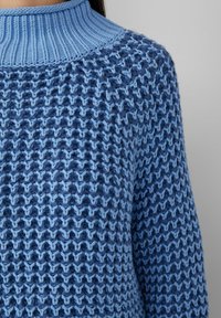 Vrouw in een blauwe chunky gebreide trui met een hoge ribbelkraag en een textuurpatroon op het lichaam en de mouwen.