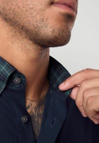 Marineblauwe shirt met een button-down kraag, voorzien van een groen en blauw geruit interieur. Inclusief twee knopen bij de kraag en een zichtbare tattoo.