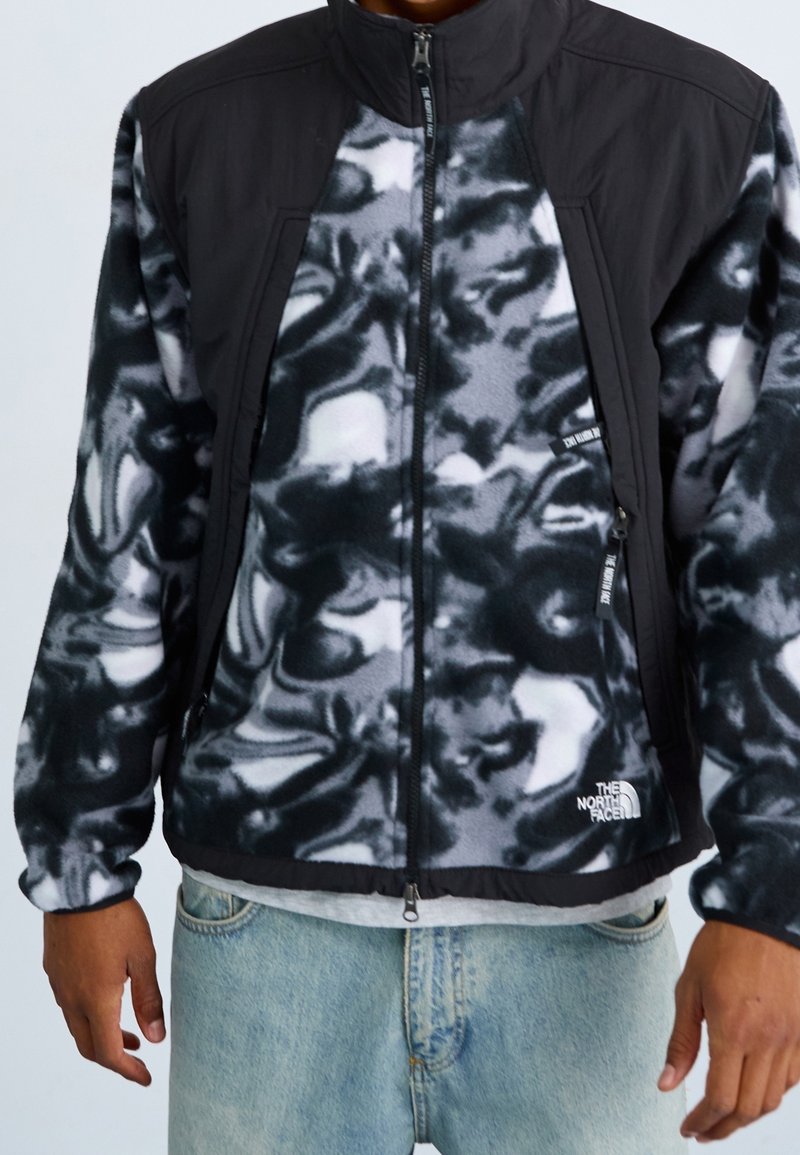 Chaqueta de forro polar con patrón abstracto en negro y gris, cierre de cremallera, cuello alto y hombros en negro sólido; presenta un logotipo en el lado izquierdo.