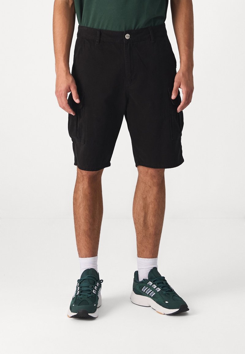 Pantaloni cargo neri con due tasche laterali, realizzati in cotone. Abbinati a scarpe sportive verdi e calzini bianchi. Design semplice con chiusura a bottone.