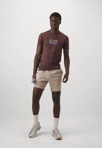 Bordeaux atletisk t-skjorte med "EA7" logo, beige shorts, sølvfargede joggesko, holder en manual. Jevn stoff, tettsittende design og synlig søm.