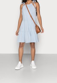 Frau trägt ein hellblaues, ärmelloses, gestuftes Kleid, weiße Sneaker und eine braune Umhängetasche vor einem einfarbigen Hintergrund.