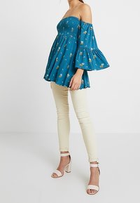 Teal paita, joka on olkapäiden alapuolelta auki, kukkakuosisena ja hieman hulmuavilla hihoilla, yhdistettynä vaaleisiin skinny-housuihin ja vaaleanpunaisiin korokepolkkaisiin.