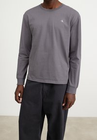 Harmaa pitkähihainen t-paita, joka on valmistettu puuvillasta. siinä on pyöreä kaula-aukko ja pieni kirjailtu logo rinnassa. Yhdistetty tummiin housuihin.