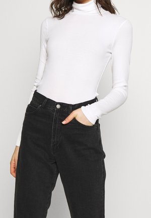 Frau trägt einen enganliegenden weißen Rollkragenpullover und hochtaillierte schwarze Jeans, eine Hand in der Tasche, vor einfarbigem Hintergrund.