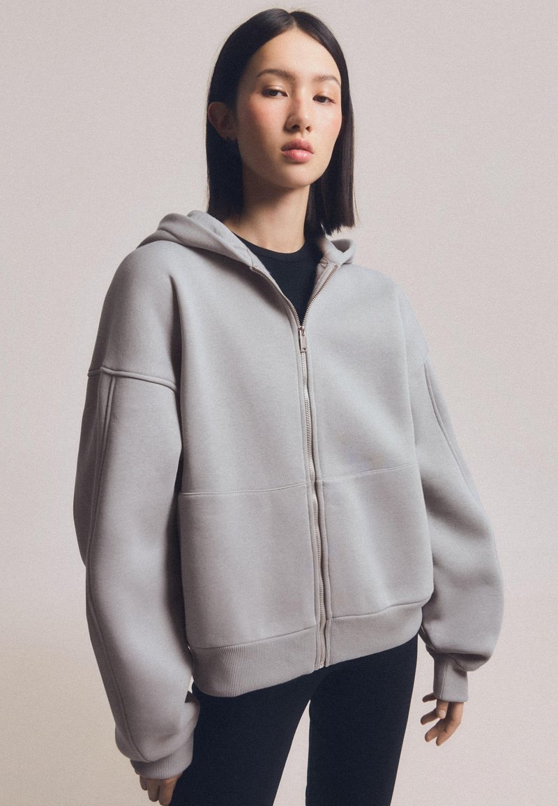 Szürke, cipzáros kapucnis pulóver, laza szabású, nagyméretű ujjakkal, bordás mandzsettával és tágas elülső zsebével, puha, strukturált anyagból készült.