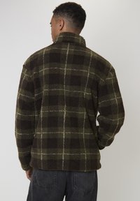 Mężczyzna w ciemnozielonej i brązowej kratkowanej polarowej kurtce, widoczny od tyłu, w połączeniu z czarnymi dżinsami.