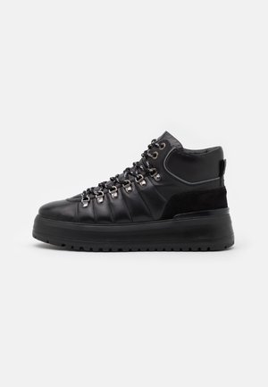 Zwarte hoge sneakers met leren en suède bovenkant, metalen veterogen, dikke rubberen zool en patroonveters, vanaf de zijkant weergegeven op een witte achtergrond.