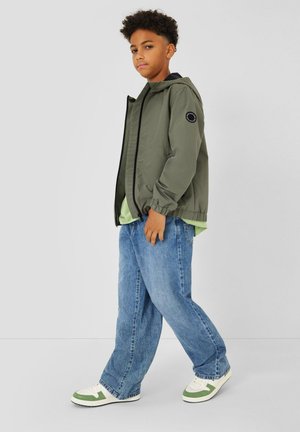 Junge mit lockigem Haar, der eine olivgrüne Kapuzenjacke, ein hellgrünes Hemd, locker sitzende blaue Jeans und weiße-grüne Turnschuhe trägt, steht seitlich.