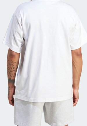 Vit t-shirt i bomull med avslappnad passform, korta ärmar och rak fåll, bärs tillsammans med ljusgrå shorts. Tatueringar syns på armen.