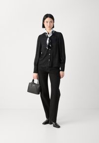 Blouse noire texturée avec de longues manches et des boutons, assortie à un pantalon noir à jambe droite et un petit sac à main noir. Finitions avec des chaussures foncées.