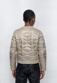 Vadderad beige kort jacka med ett vågmönster och en slät yta, ihopparad med svarta läderbyxor. Enhetlig bakgrund.