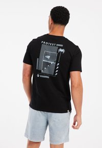 Schwarzes Baumwoll-T-Shirt mit grafischem Druck in hellblau auf dem Rücken, mit geometrischen Mustern und Text, getragen zu hellblauen Shorts.