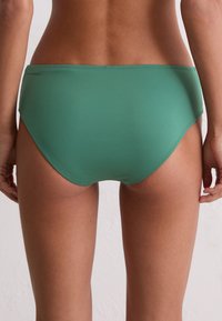 Kobieta z odkrytą dolną częścią pleców i biodrami w zielonych, bezszwowych majtkach na tle neutralnej ściany.