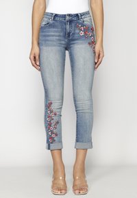 Hellblaue Denim-Jeans mit floralen Stickereien in Rot und Weiß am linken Bein, schlank geschnitten mit hochgekrempelten Säumen am Knöchel.