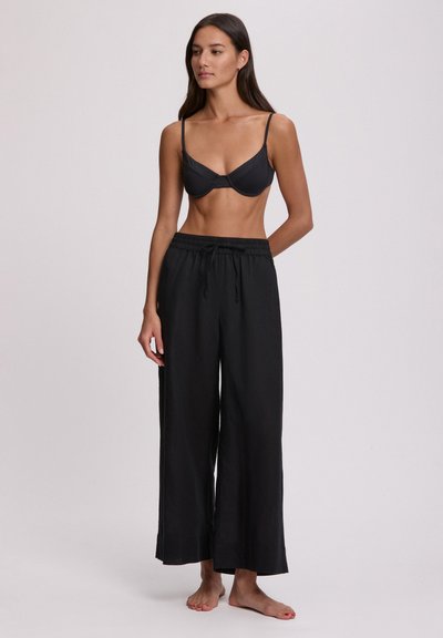 Γυναίκα με μακριά σκούρα μαλλιά, φορώντας μαύρο σουτιέν και άνετο μαύρο παντελόνι, όρθια ξυπόλυτη μπροστά σε απλό φωτεινό φόντο.