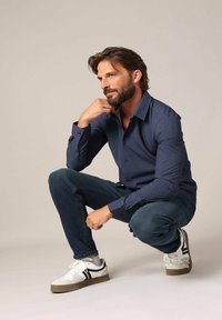 Marineblauwe shirt met een klein geometrisch patroon, gecombineerd met donkere jeans en witte sneakers met zwarte accenten. Strak geweven stof.