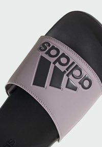 Fialová šmykľavá sandála s širokým pásikovým dizajnom, zvýrazneným čiernym logom a textúrovanou čiernou podrážkou, zdôrazňujúcou pohodlie a priľnavosť.