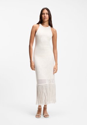 Femeie purtând o rochie maxi tricotată albă fără mâneci, cu tiv franjurat și tocuri albe decupate la degete, stând în picioare pe un fundal simplu.
