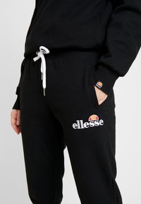 Svarta sweatpants i bomull med vit dragsko och elastisk midja. Har en vit "ellesse"-logotyp och en liten färgad logotyp på fickan.