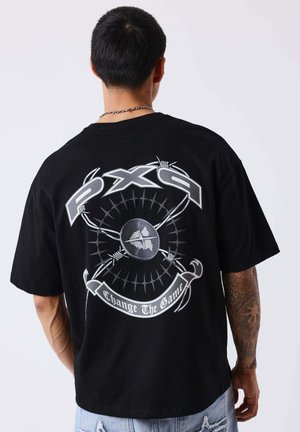 Homme avec un bras tatoué portant un t-shirt noir avec un "PX9" argenté et un graphisme de barbelés ainsi qu'un texte "Change The Game" dans le dos.