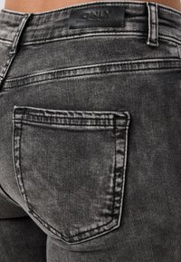 Vaqueros de mezclilla negra con un acabado desgastado, diseño de cinco bolsillos y una etiqueta de cuero con la marca en la cinturilla. Vista en primer plano.