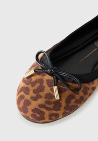 Ballerine en suède à imprimé léopard marron et noir avec un nœud en ruban noir. Présente des accents dorés sur les extrémités du nœud. Design à bout arrondi.