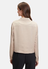 Lichtbeige cropped jacket met een kraag, lange mouwen en knoopmanchetten; gladde textuur met een subtiel geweven patroon.
