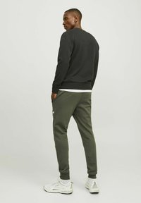 Zwarte crewneck sweatshirt gecombineerd met olijfgroene joggers. Beide kledingstukken zijn gemaakt van stof met een gestructureerde afwerking. Witte sneakers maken de outfit compleet.