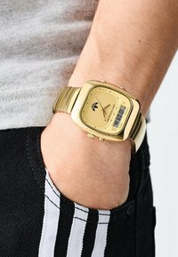 Guld digital klocka med fyrkantig urtavla, en texturerad gul metallarmband och svarta accenter, som visar tiden i digitalt format.