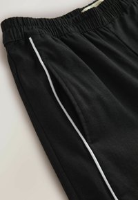 Pantalon avec taille élastique noire, orné d'un détail de passepoil blanc le long de la couture latérale et de l'ouverture de la poche avant.
