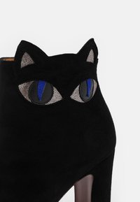 Bottine noire en daim à talon haut avec oreilles de chat et yeux de chat brodés en gris, noir et bleu sur le devant.
