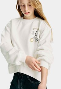 Sweat-shirt blanc avec un col rond, des poignets et un ourlet côtelés. Dispose d'un graphique brodé avec un porte-clés et du texte sur la poitrine.