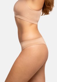 Nude bezszwowy biustonosz braletka i zestaw stringów, elastyczny materiał, prosty design, szerokie paski, miękka tekstura, ujęcie z boku modelki.