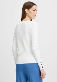 Haut à manches longues blanc à côtes avec un large col, une coupe fitted et des accents de boutons noirs sur les manches. Porté avec un pantalon bleu.