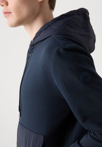 Navy blue hoodie featuring a soft fabric, adjustable drawstring hood, and a textured lower pocket; includes a circular logo on the sleeve.