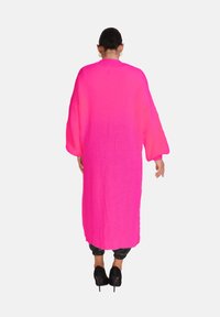 Lång, löst sittande, stickad kappa i klar rosa. Har vida ärmar, en strukturerad tyg och är stylad över svarta, figursydda byxor och klackar.